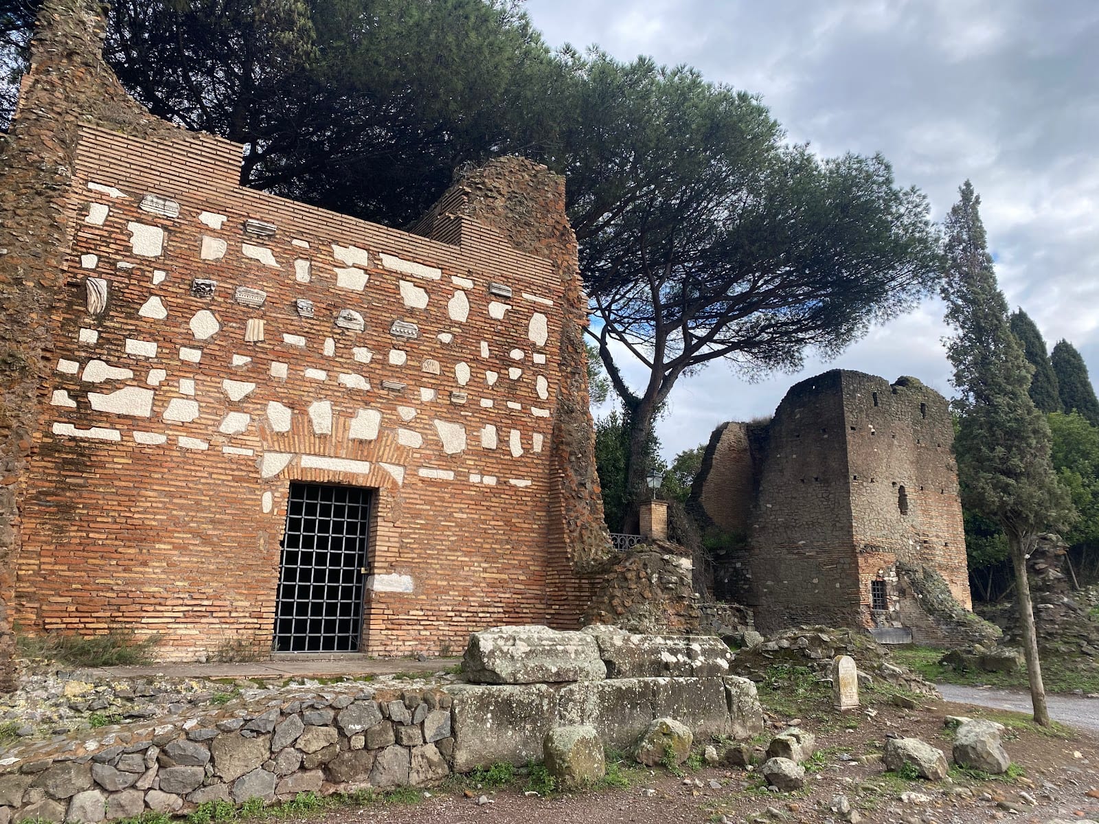 Ruined Roman tombs and an umbrella pine along the basalt cobblestones of the Appian Way under an autumn sky.