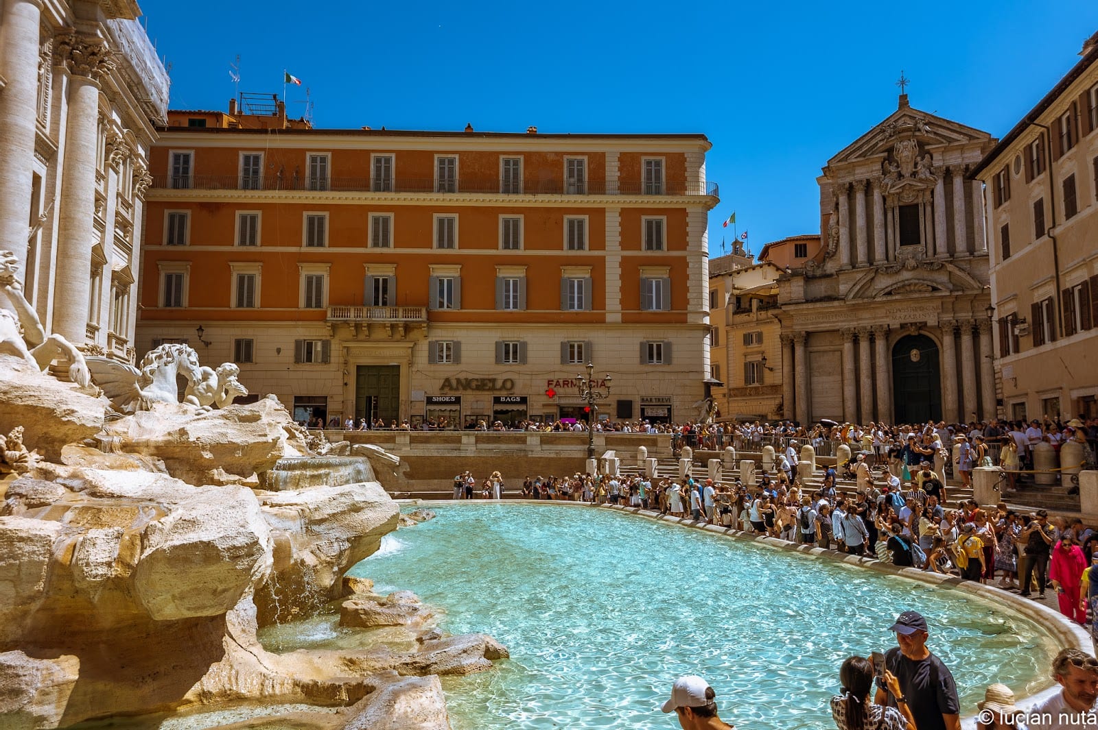Crowds around the turquoise basin of the Trevi Fountain by day.