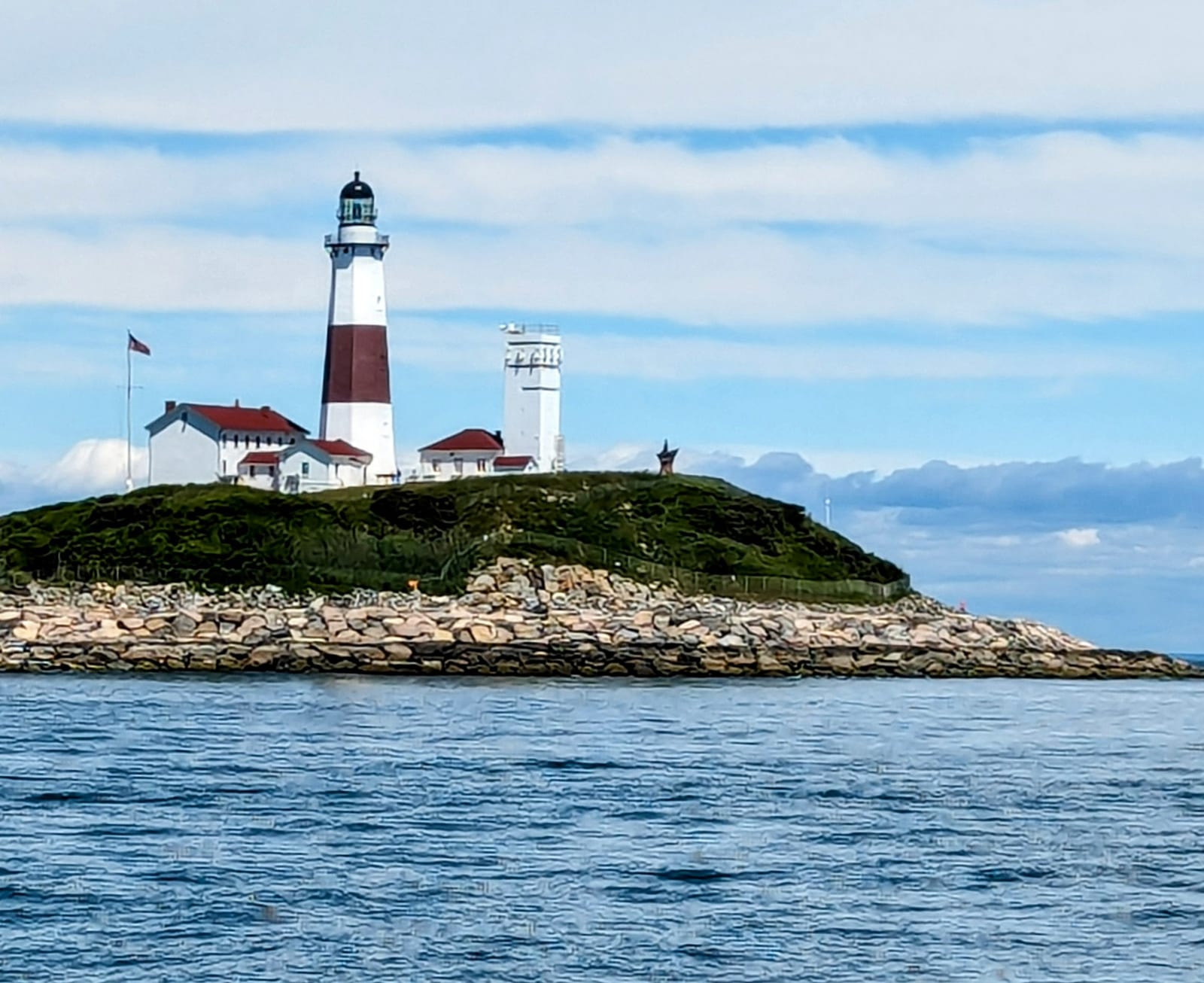 Montauk Point Lighthouse and its surrounding buildings stand on a rocky shore above the water.