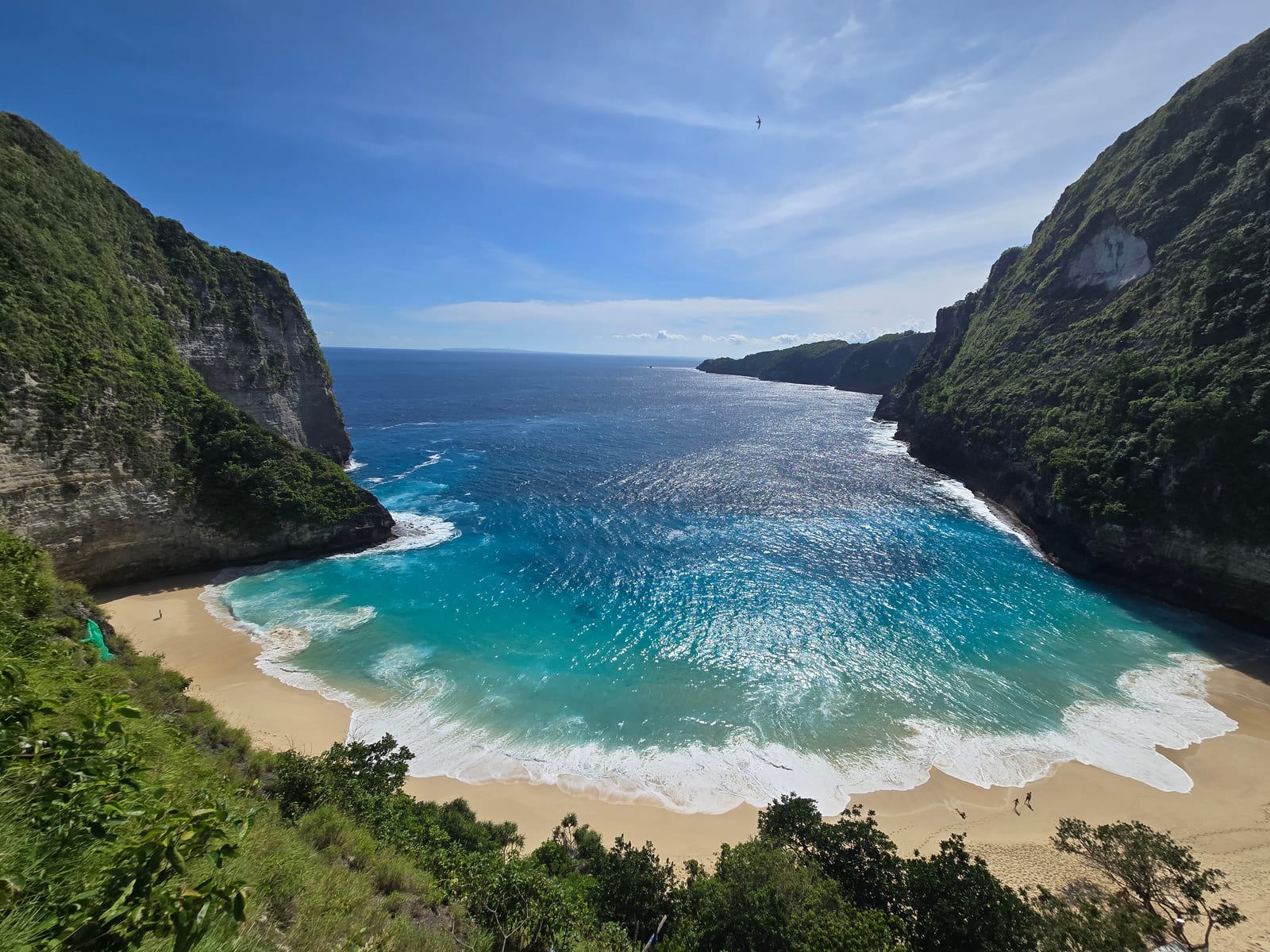 Kelingking Beach lies below steep green cliffs, with a rocky headland and turquoise waves along the sandy shore.