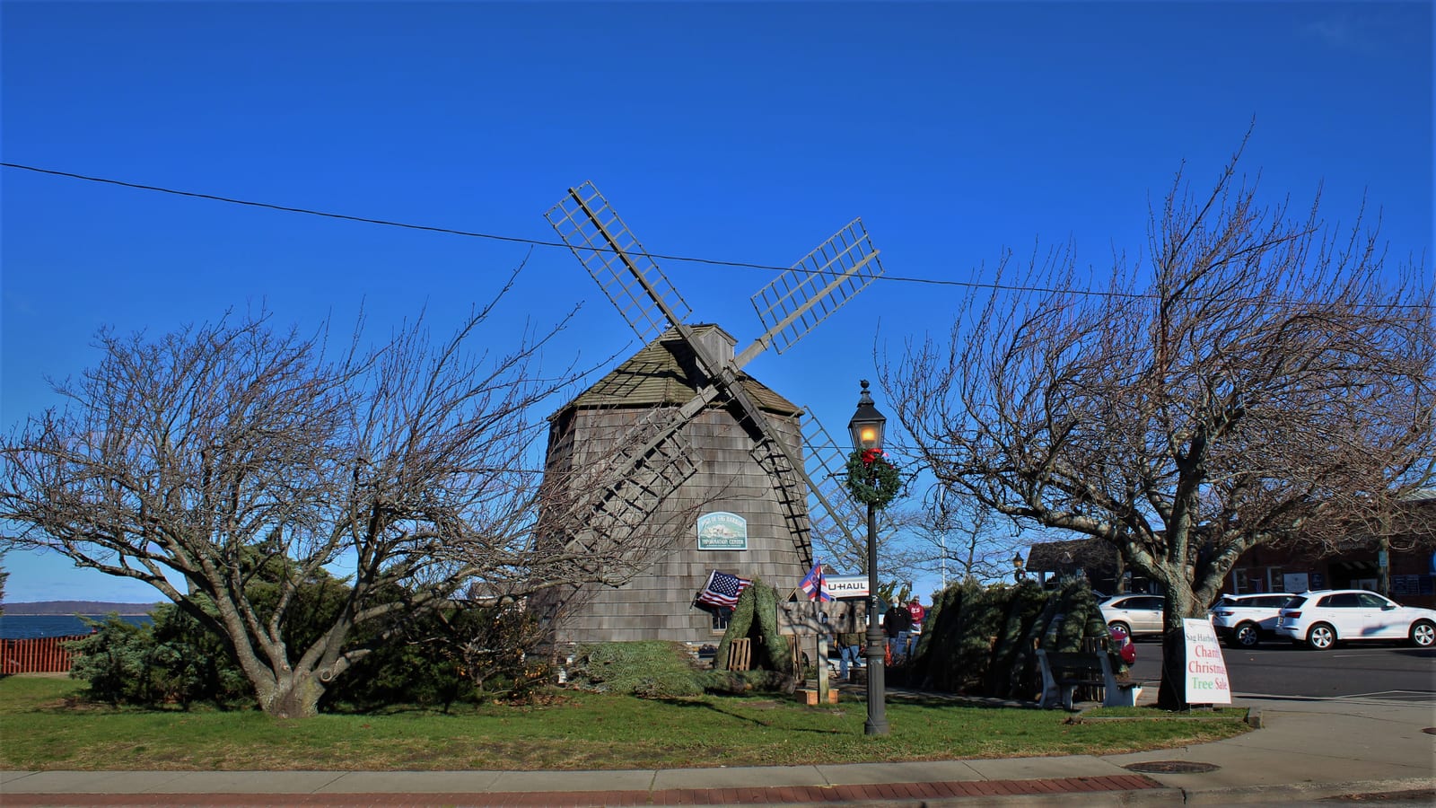 The Sag Harbor Windmill stands among leafless trees beside a street under a clear blue sky.