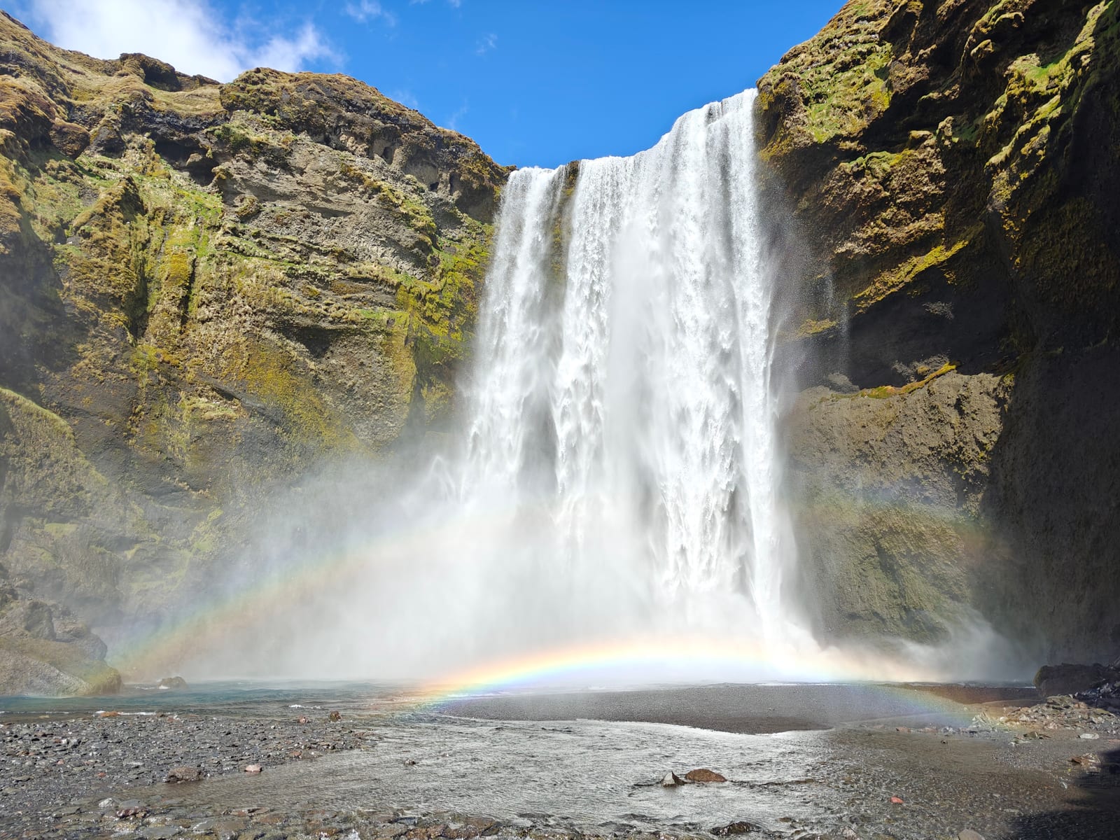 Skogafoss drops over a moss-covered cliff beneath a bright blue sky.