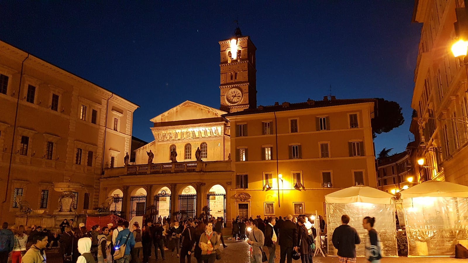 The lit basilica and crowd on Piazza di Santa Maria in Trastevere at night.