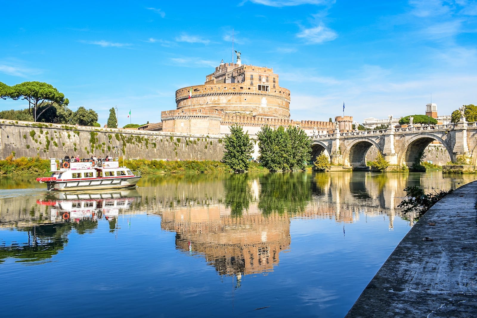 Castel Sant'Angelo and its bridge reflected in the Tiber under a blue sky.