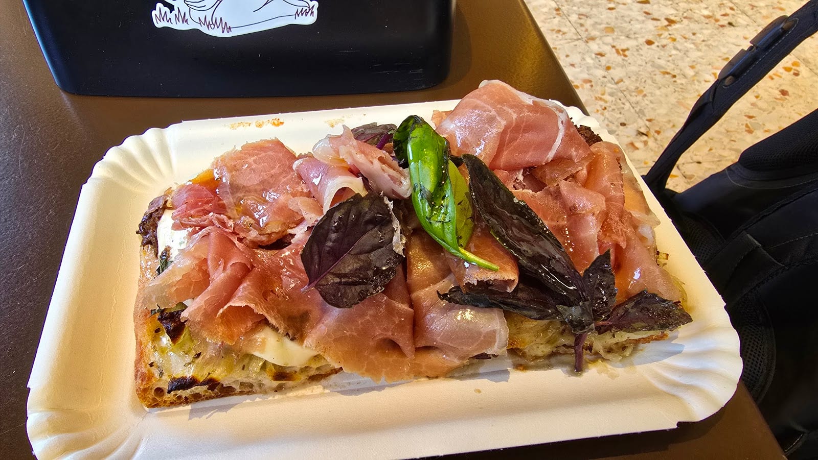 A slice of Pizzarium pizza topped with prosciutto and basil.