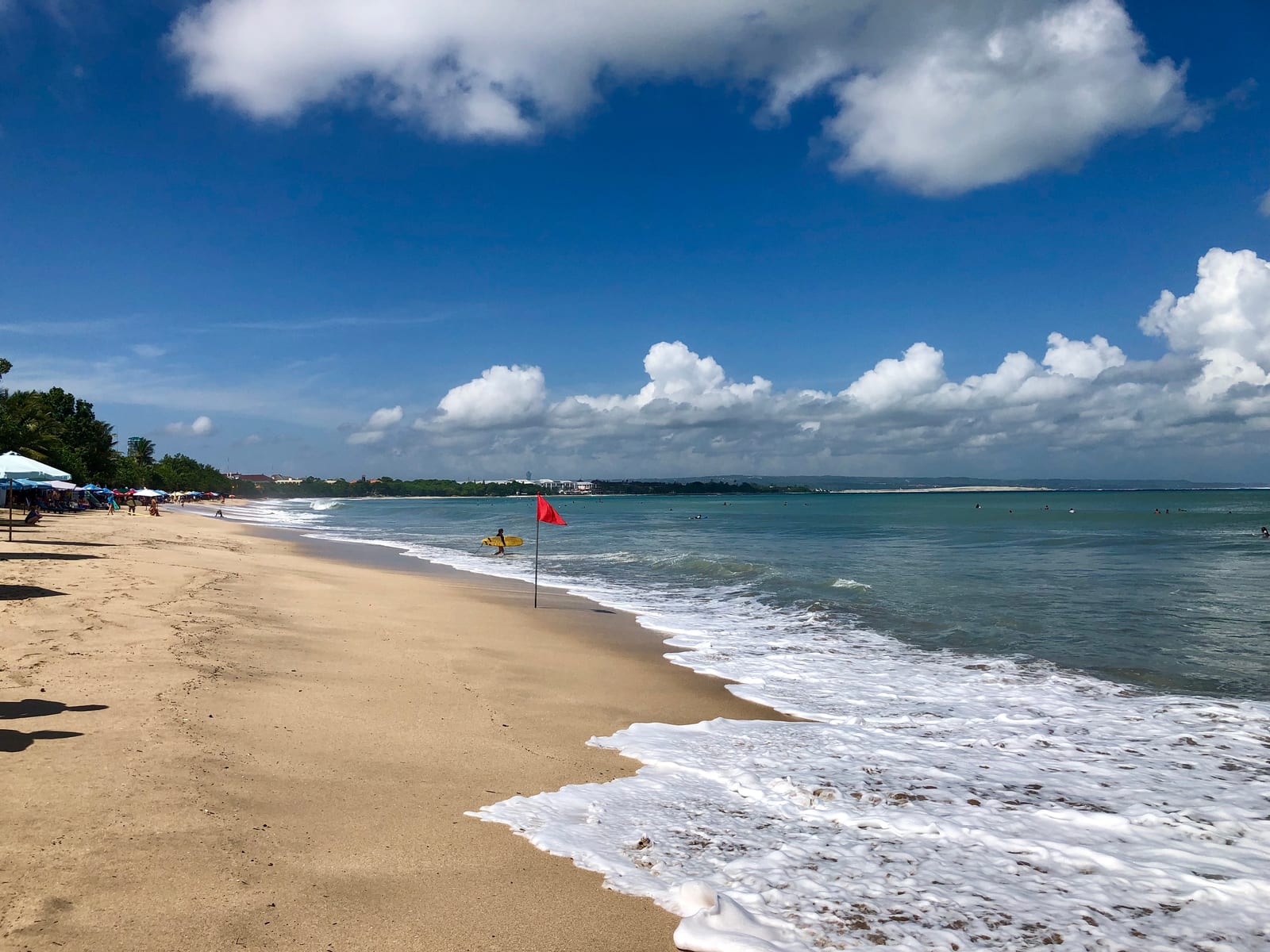 A sandy beach curves along blue water with waves, umbrellas, a red warning flag, and a surfer carrying a board.