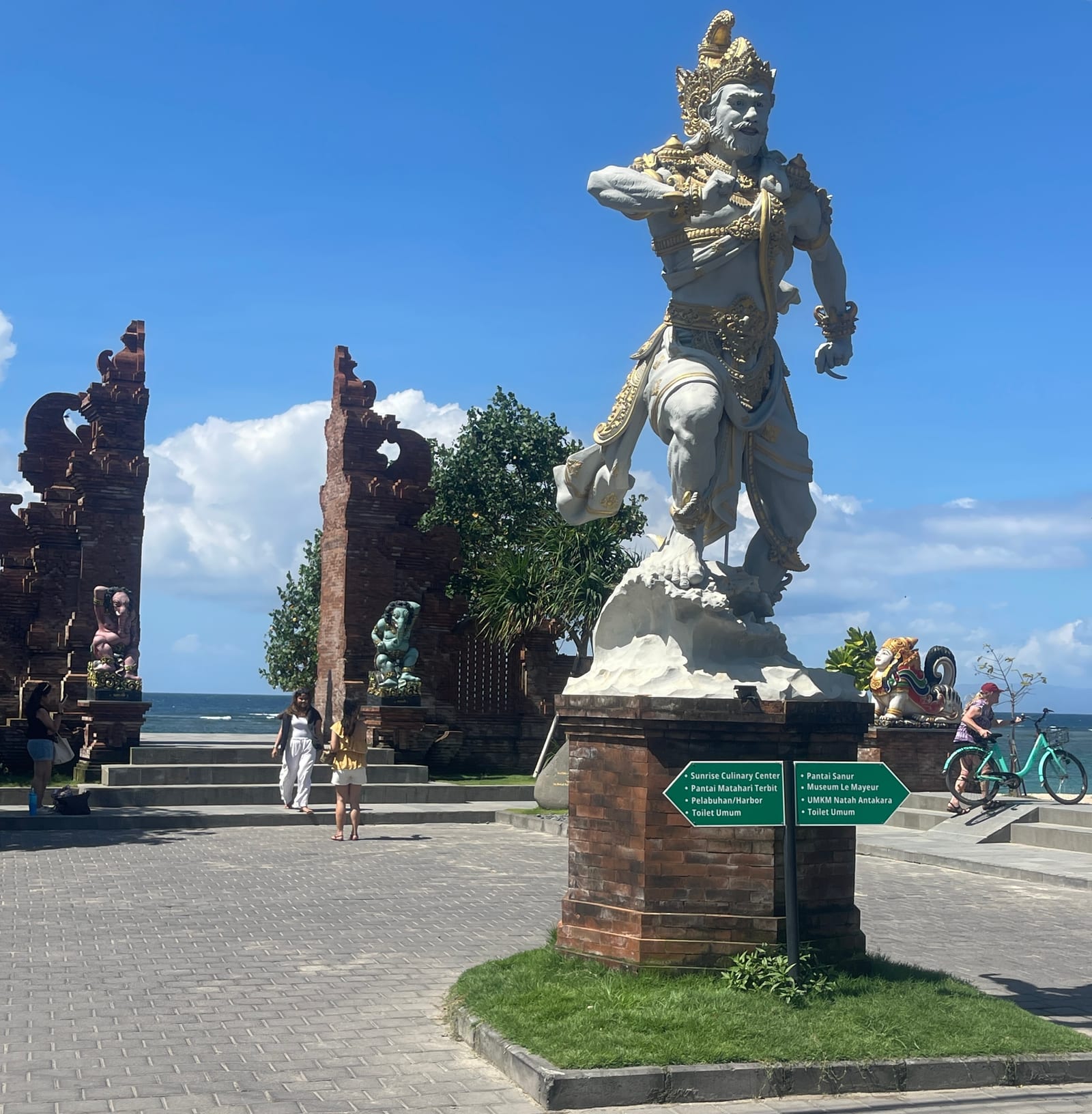 A large Balinese statue and temple gates stand beside Sanur Beach, with the sea visible behind them.
