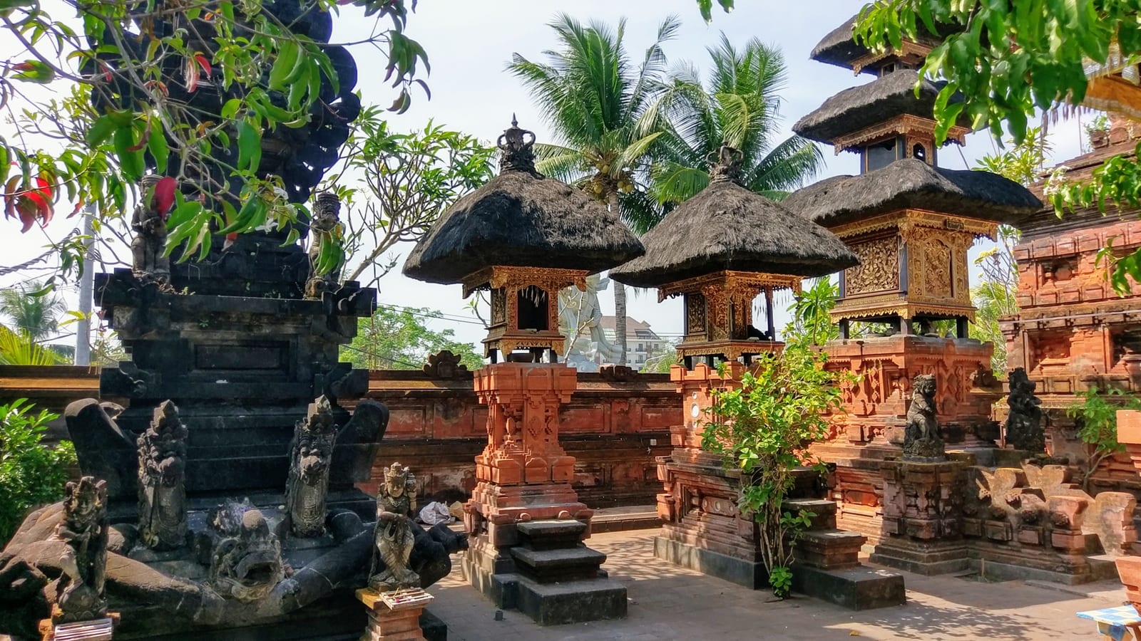 A courtyard inside Petitenget Temple with brick shrines, thatched-roof pavilions, carved stone figures, and tropical vegetation.
