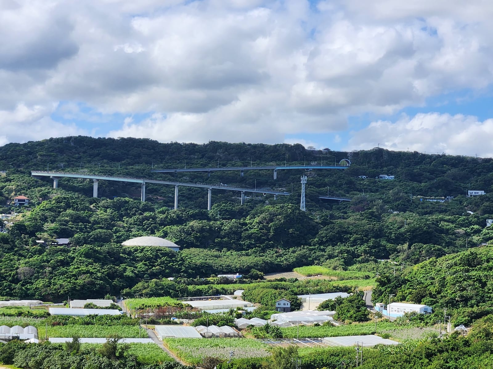 Nirai Kanai Bridge spans a green hillside above the coast, with fields and roads in the foreground.