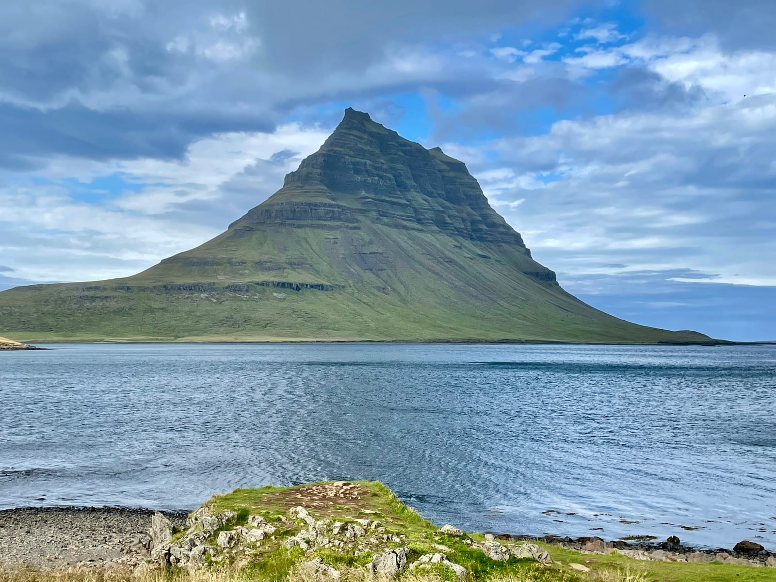 Kirkjufell rises above the water with its layered slopes and pointed summit beneath a partly cloudy sky.