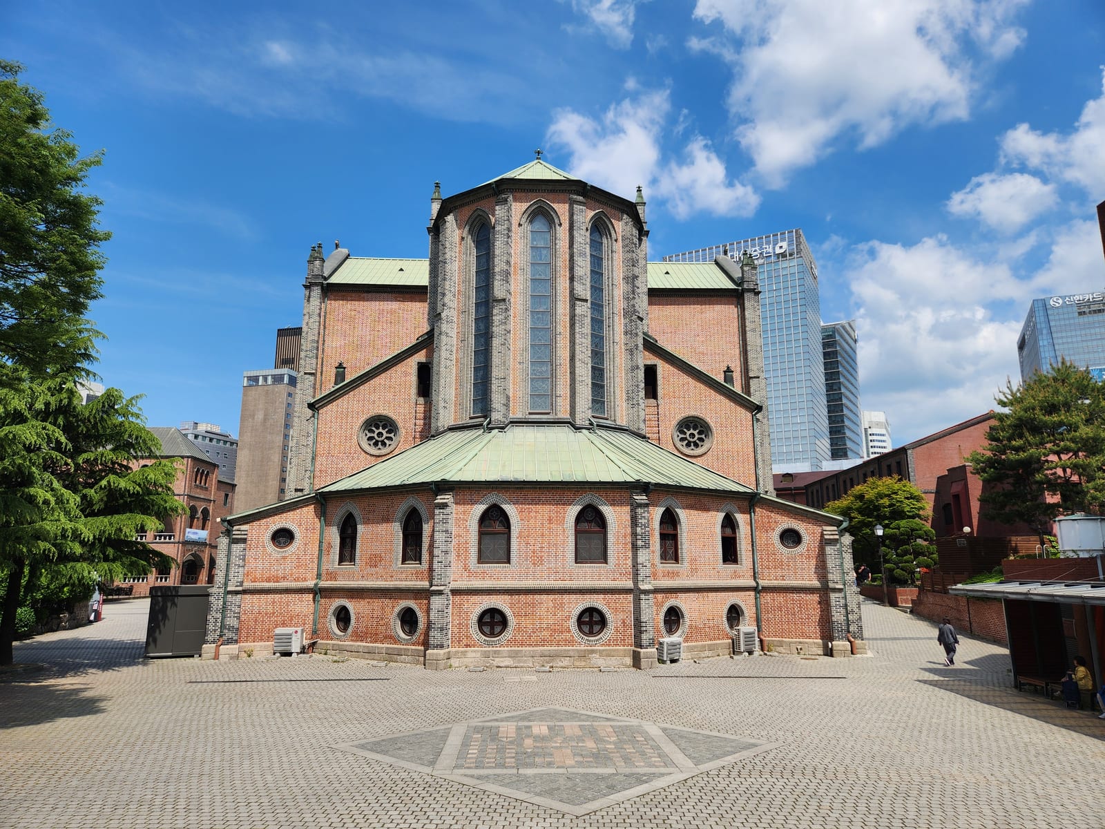 The red-brick Myeongdong Cathedral is shown from the courtyard with green roofs, trees, and a blue sky.