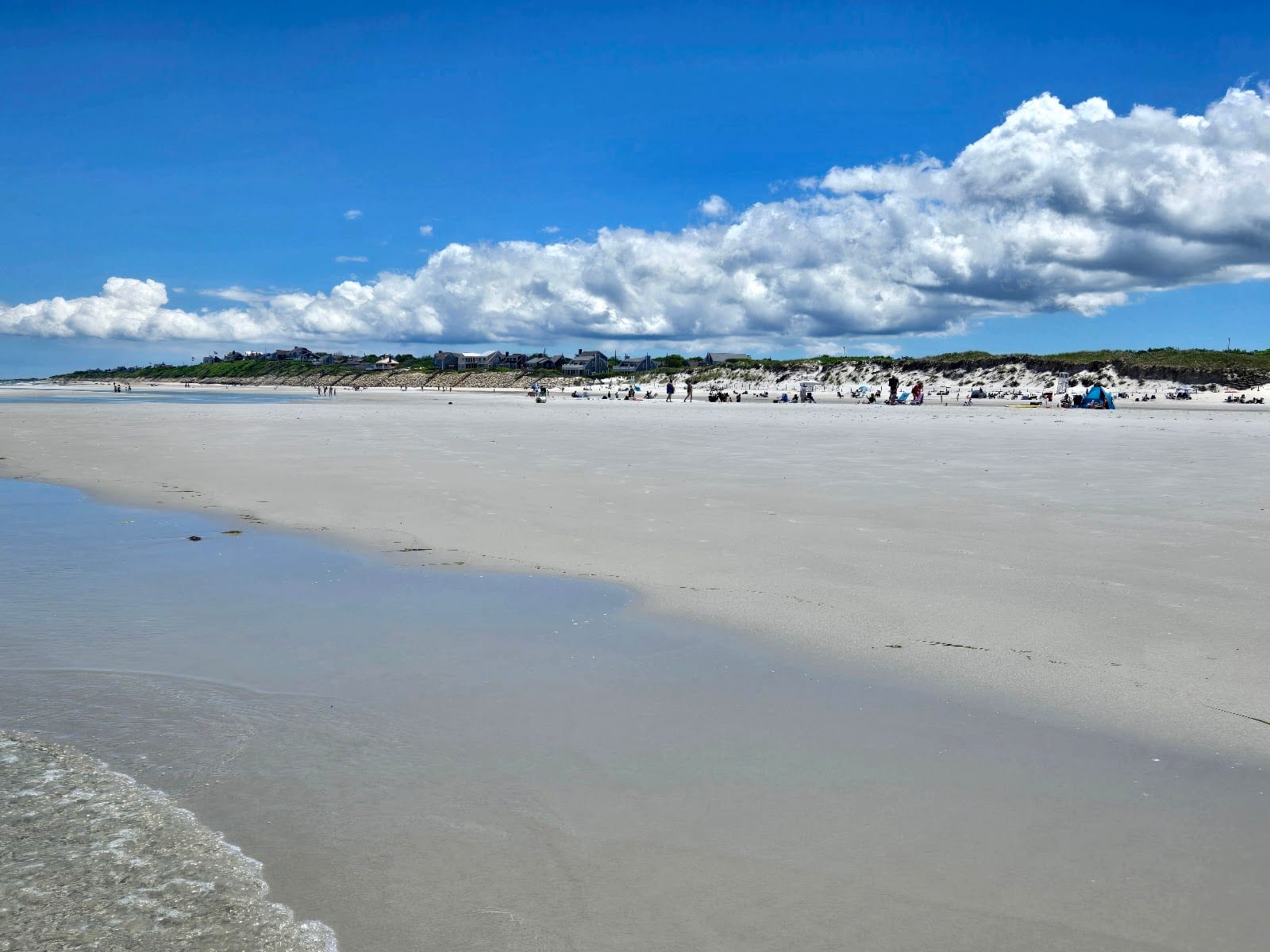Beachgoers spread across the wide low-tide flats at Mayflower Beach beneath puffy clouds.