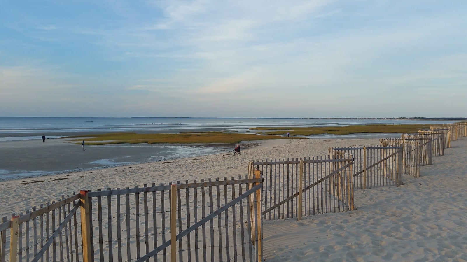 A dune fence beside the wide low-tide flats at Skaket Beach under a bright sky.