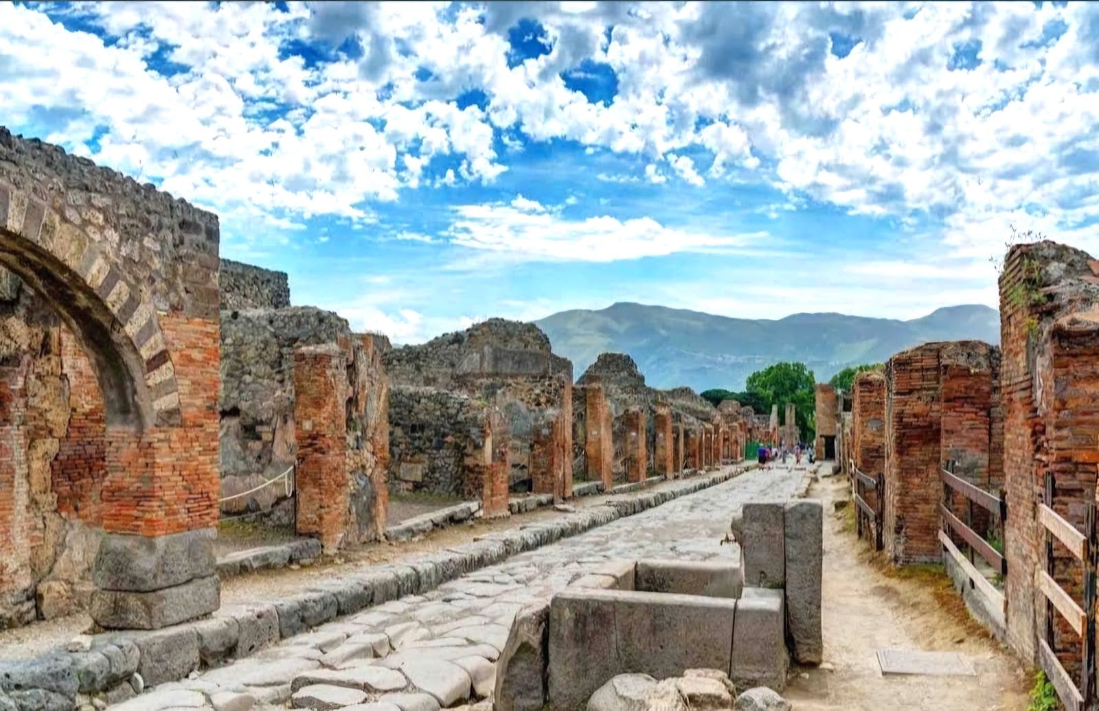 A paved ancient street of Pompeii lined with brick ruins, mountains in the distance.