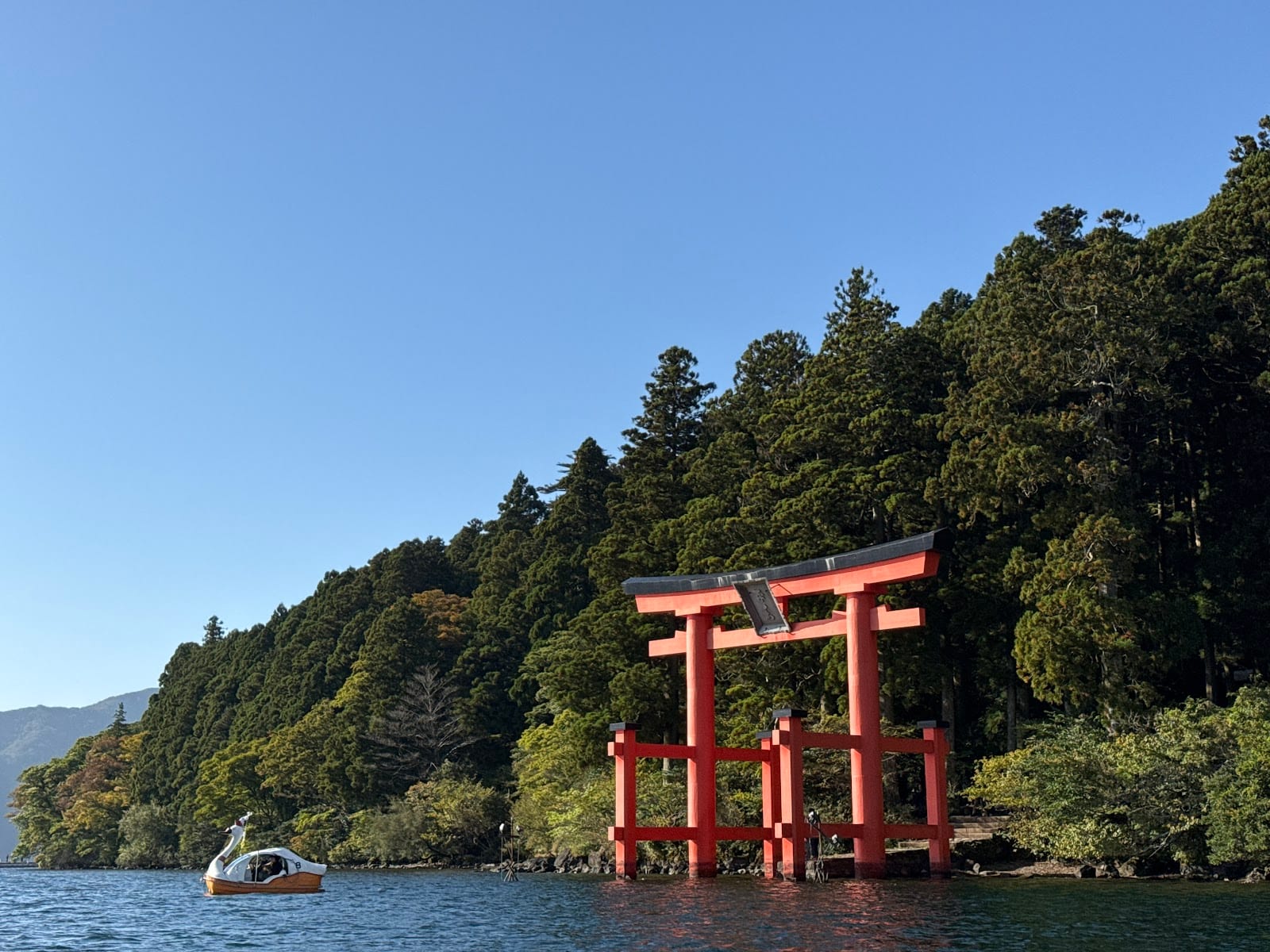 A red sightseeing galleon and the vermilion Heiwa-no-Torii of Hakone Shrine on Lake Ashi, with hills beyond.