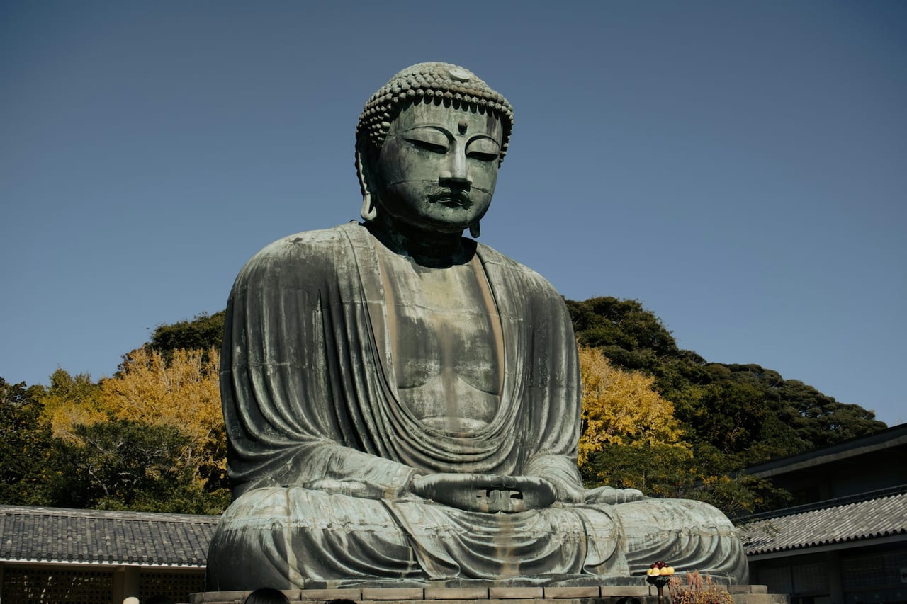 The Great Buddha of Kamakura, a giant seated bronze, sits in the open air against blue sky and autumn trees.