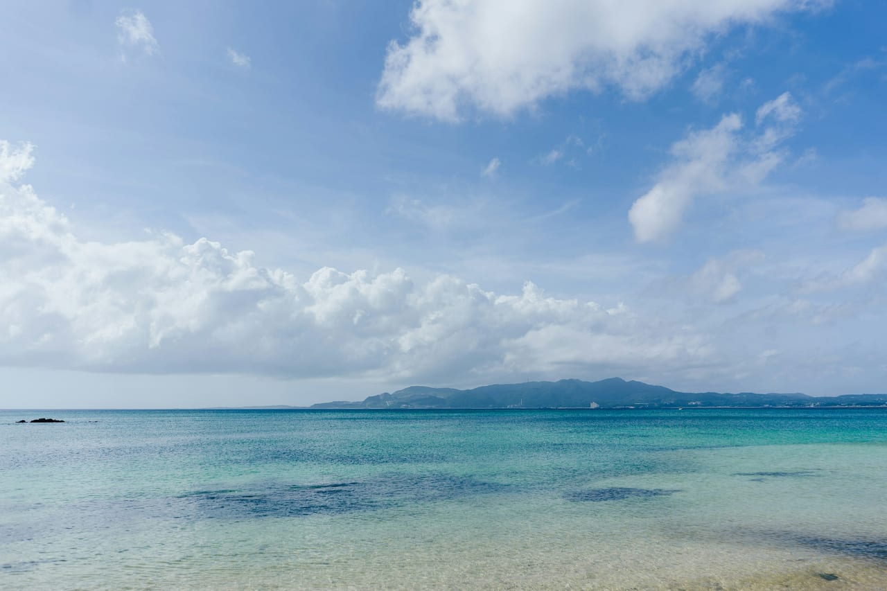 Shallow clear water and a distant mountainous coastline under scattered clouds.