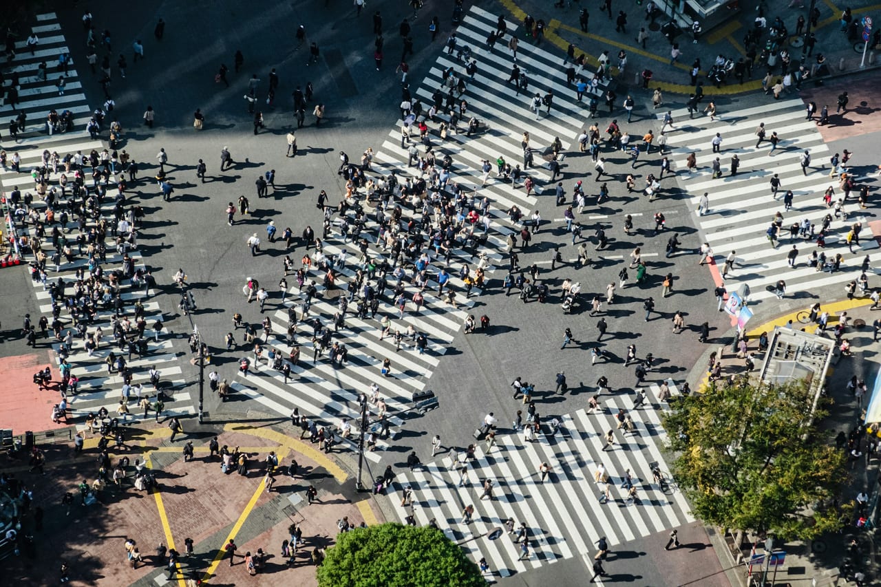 An overhead view of pedestrians flooding the diagonal lanes of the Shibuya scramble crossing in daylight.