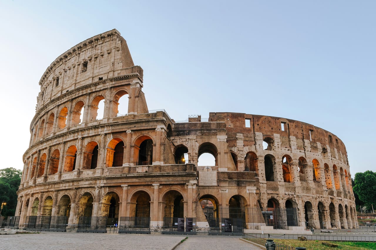 The full oval of the Colosseum stands in early light, its tiers of arches glowing gold against a pale morning sky.