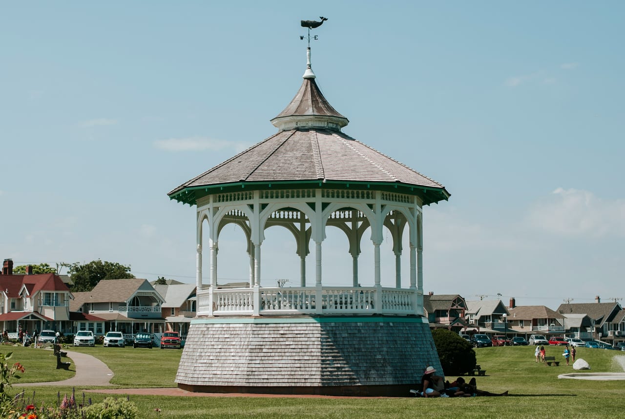 A white ornamental gazebo stands on a grassy field with houses in the background.
