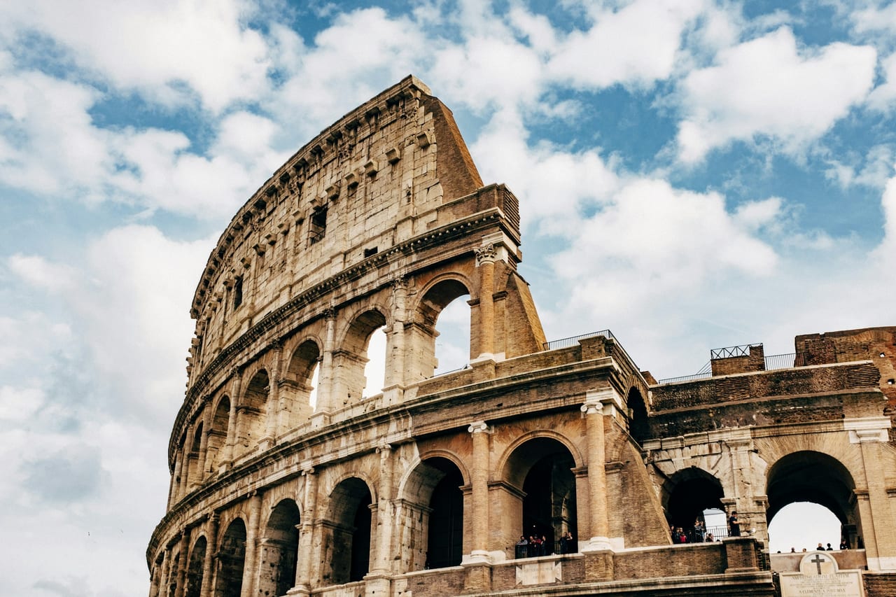 The Colosseum's weathered arches rise on a three-quarter view against a blue sky streaked with cloud.