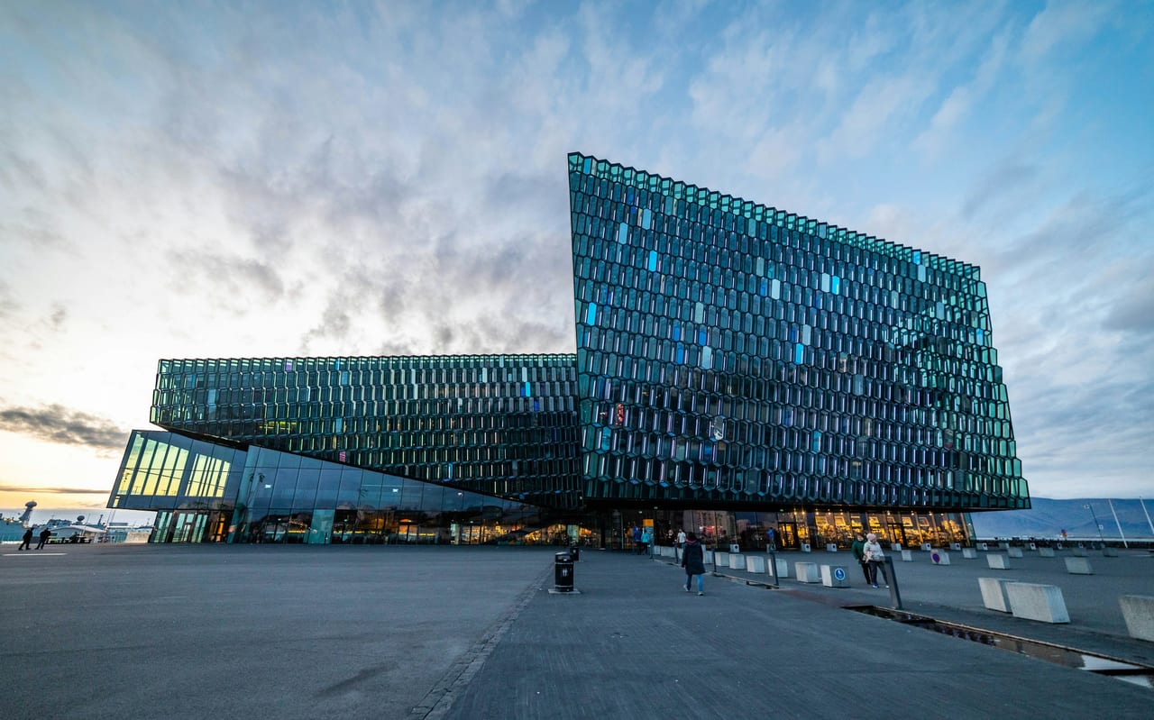 The glass Harpa concert hall stands beside Reykjavik harbor beneath a partly cloudy sky.