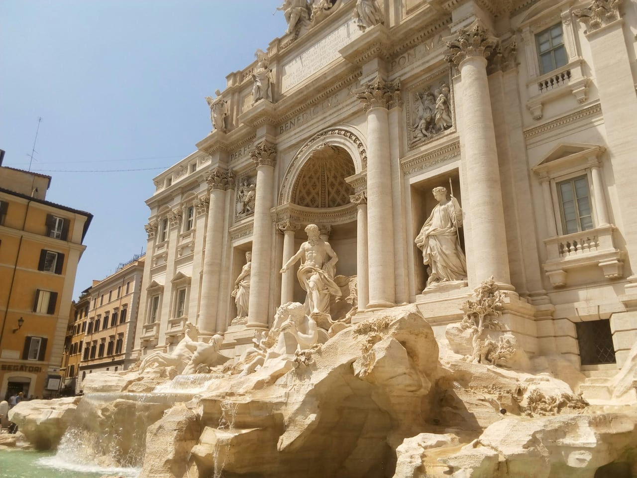 The white marble facade and sculpture of the Trevi Fountain rise against a bright blue sky above the fountain's edge.
