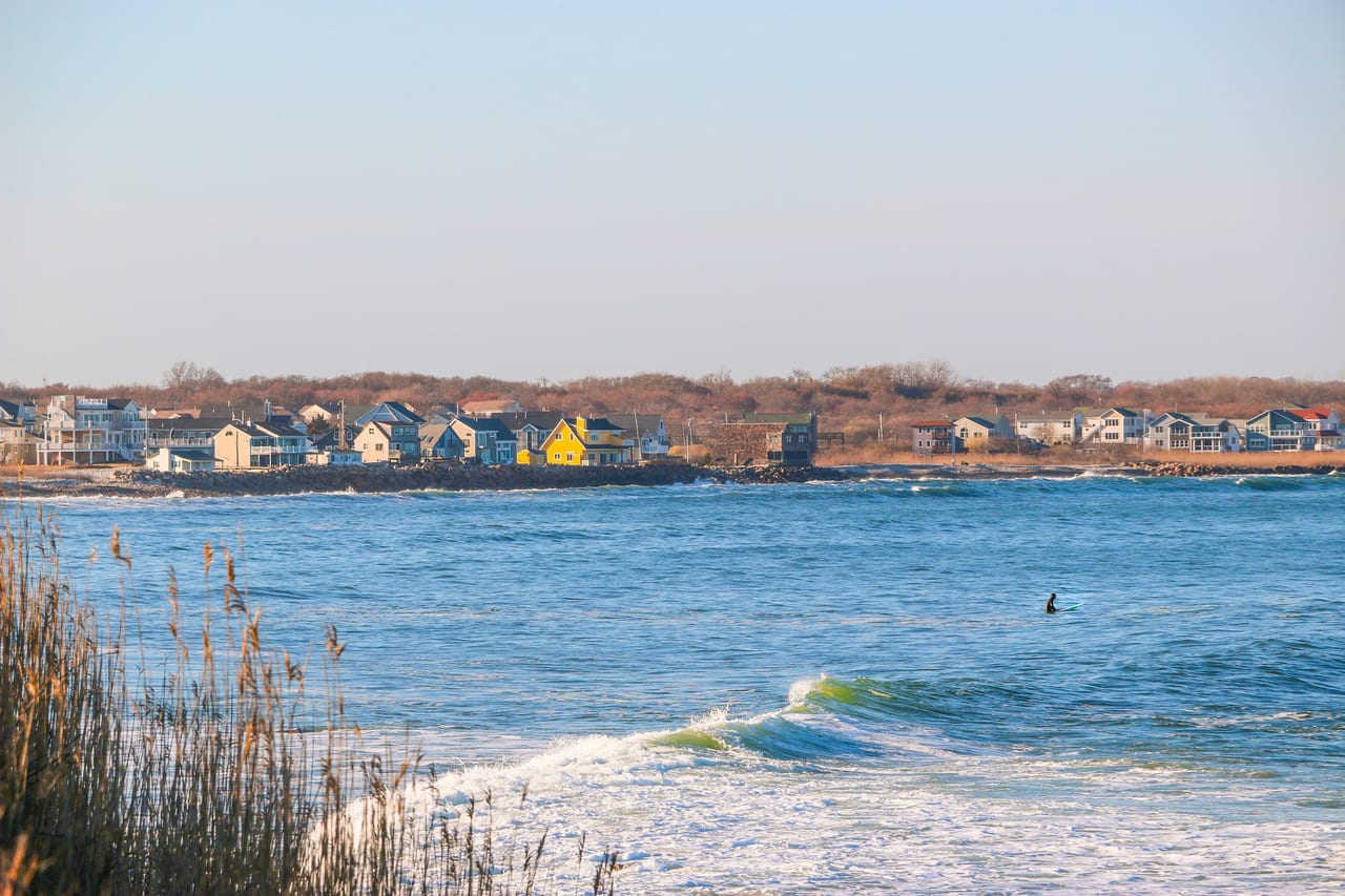 Waves break along a sandy shoreline with a row of coastal homes visible across the water on a sunny day.