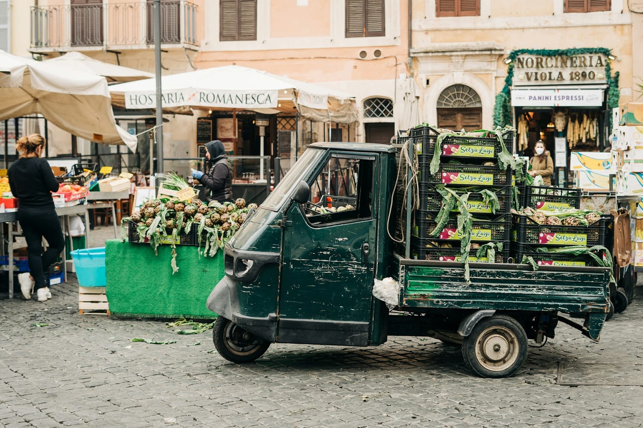 A green three-wheeled Ape van loaded with produce crates stands among the stalls of the Campo de' Fiori market.