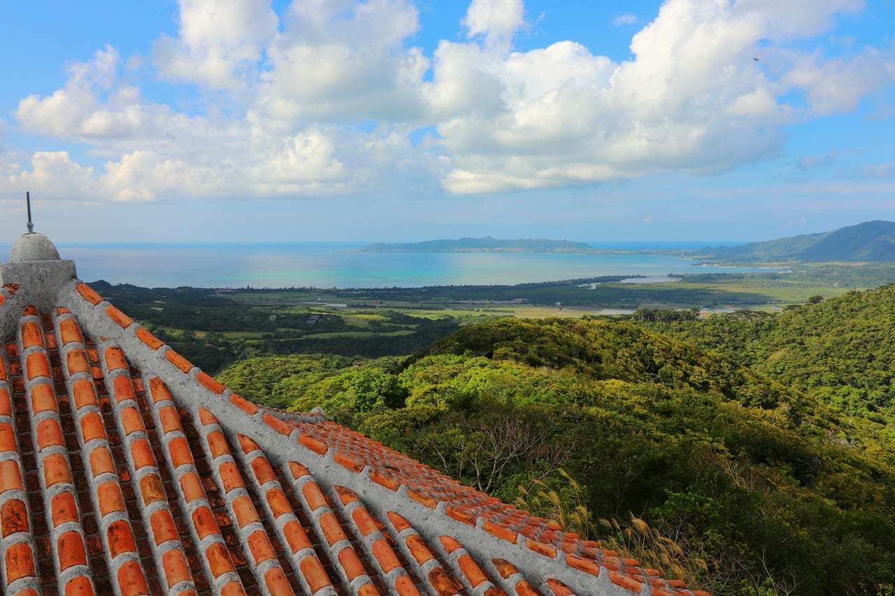 A red-tiled Okinawan roof overlooks green hills and a blue coastal bay.