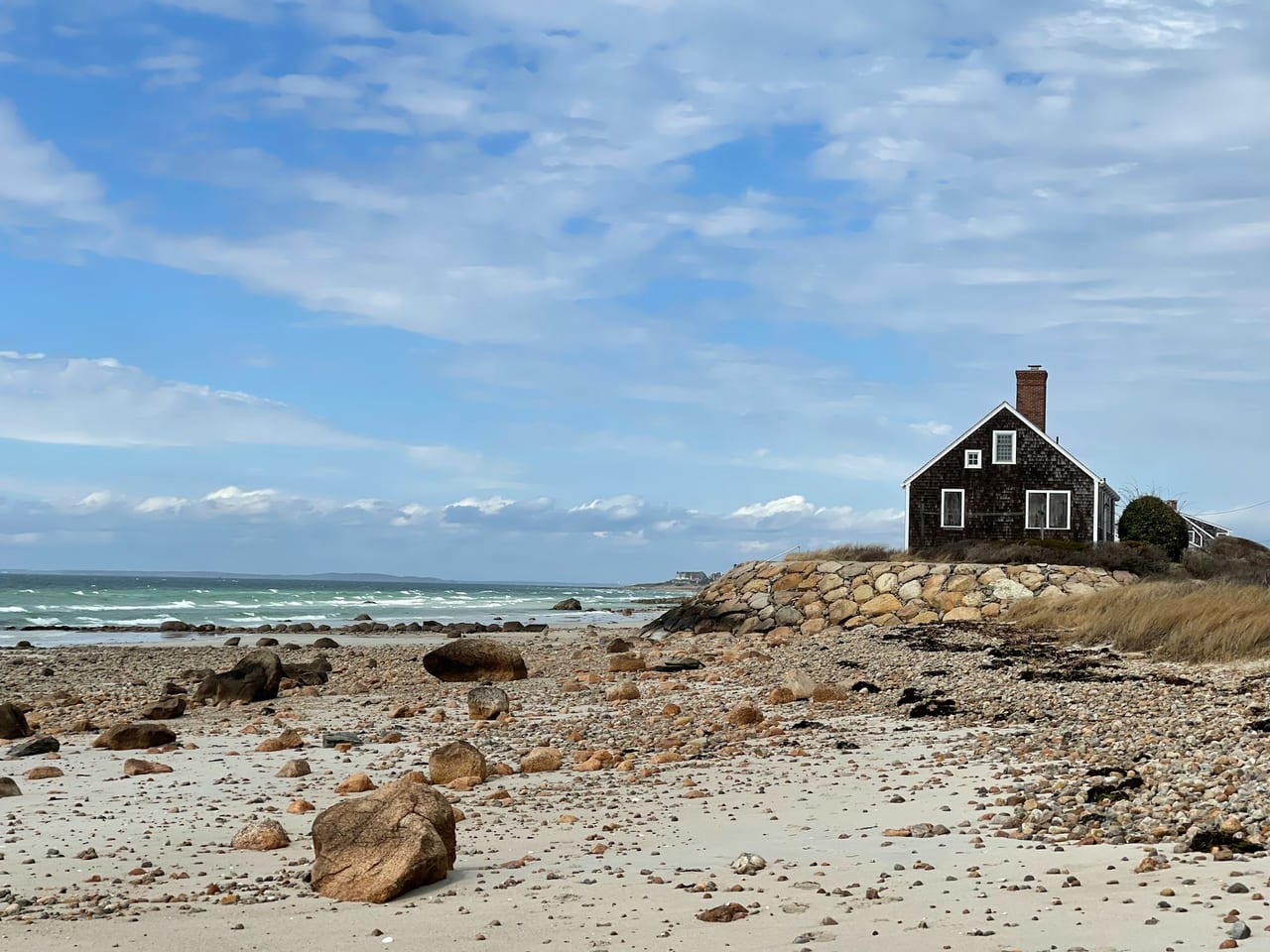 A dark shingled beach house sits above a rocky shoreline with waves rolling in under a partly cloudy blue sky.
