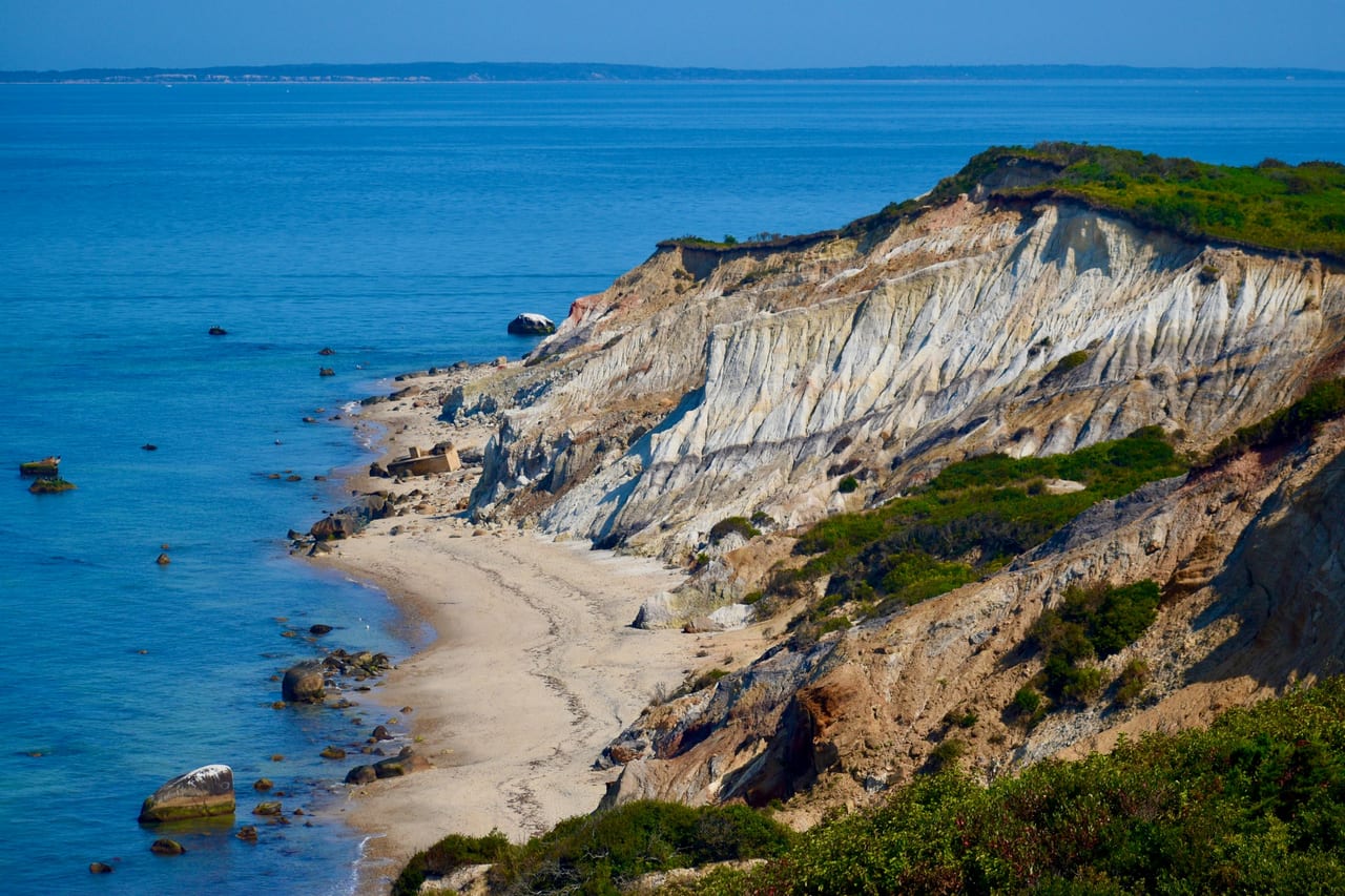 Layered clay cliffs rise above a sandy beach and clear blue ocean water under a bright sky.