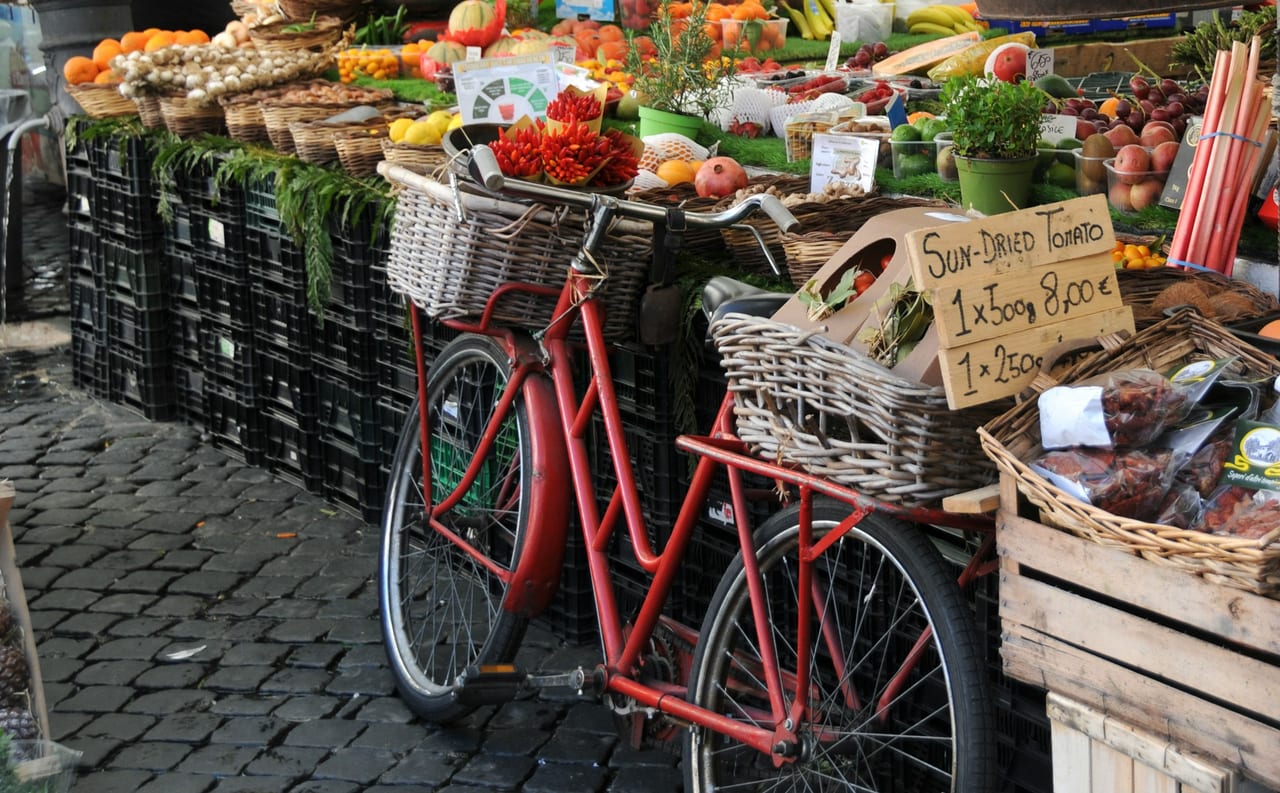 A red bicycle loaded with produce baskets leans by the laden stalls of a Rome food market.