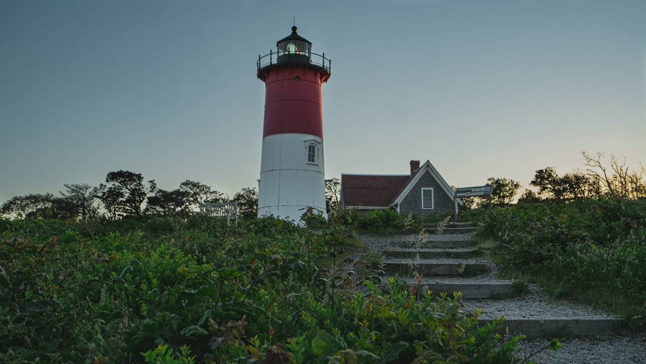 A red-and-white lighthouse stands beside a shingled keeper's house at the top of a stone-step path lined with beach shrubs.