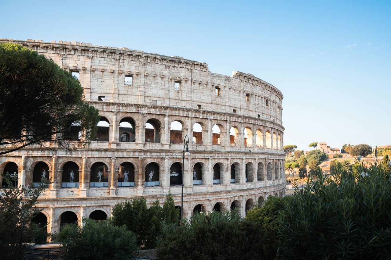 The Colosseum's tiered stone arches stand against a clear blue sky, framed by green trees.