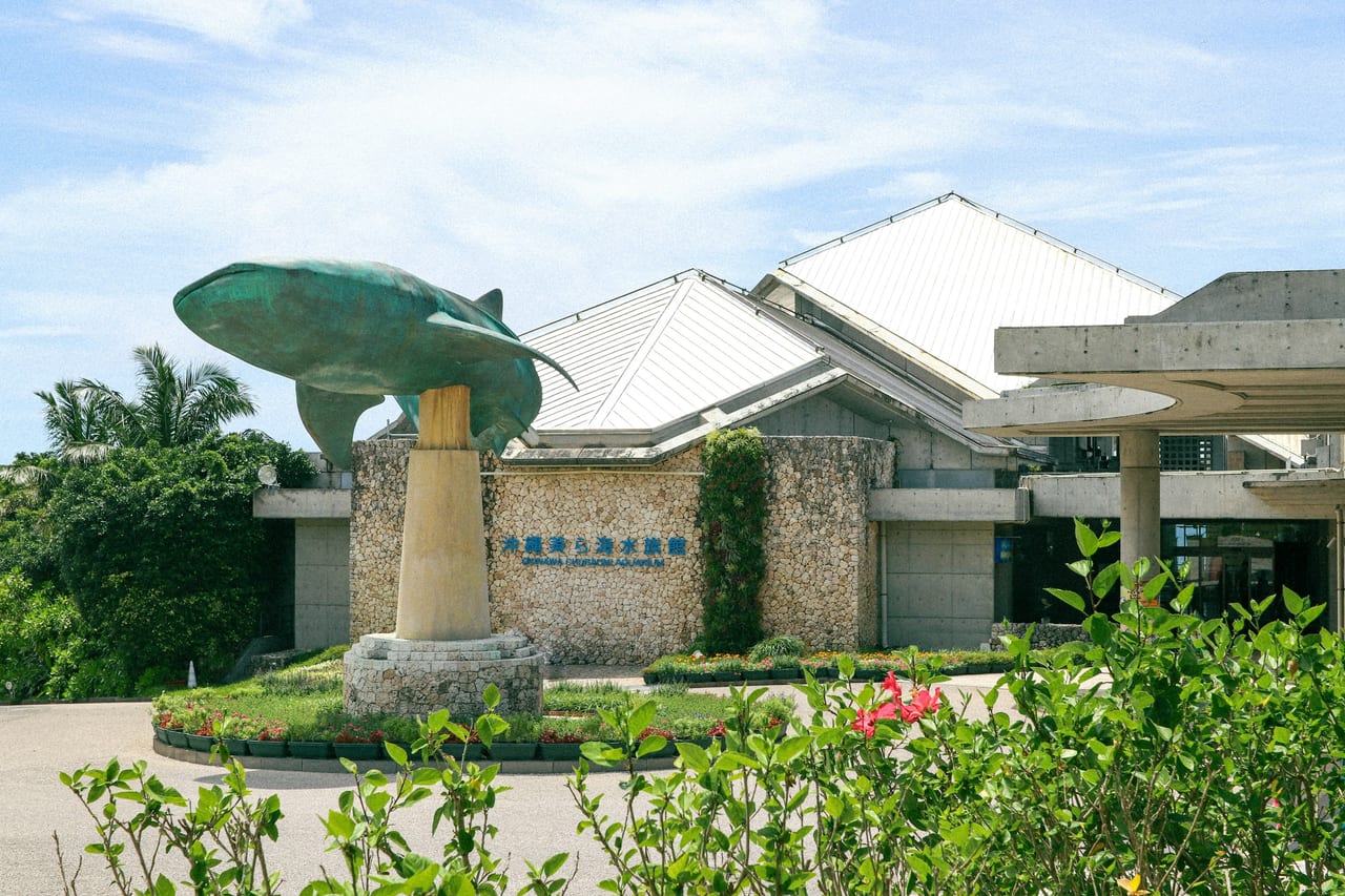 A fish statue in front of a low stone building and tropical vegetation.