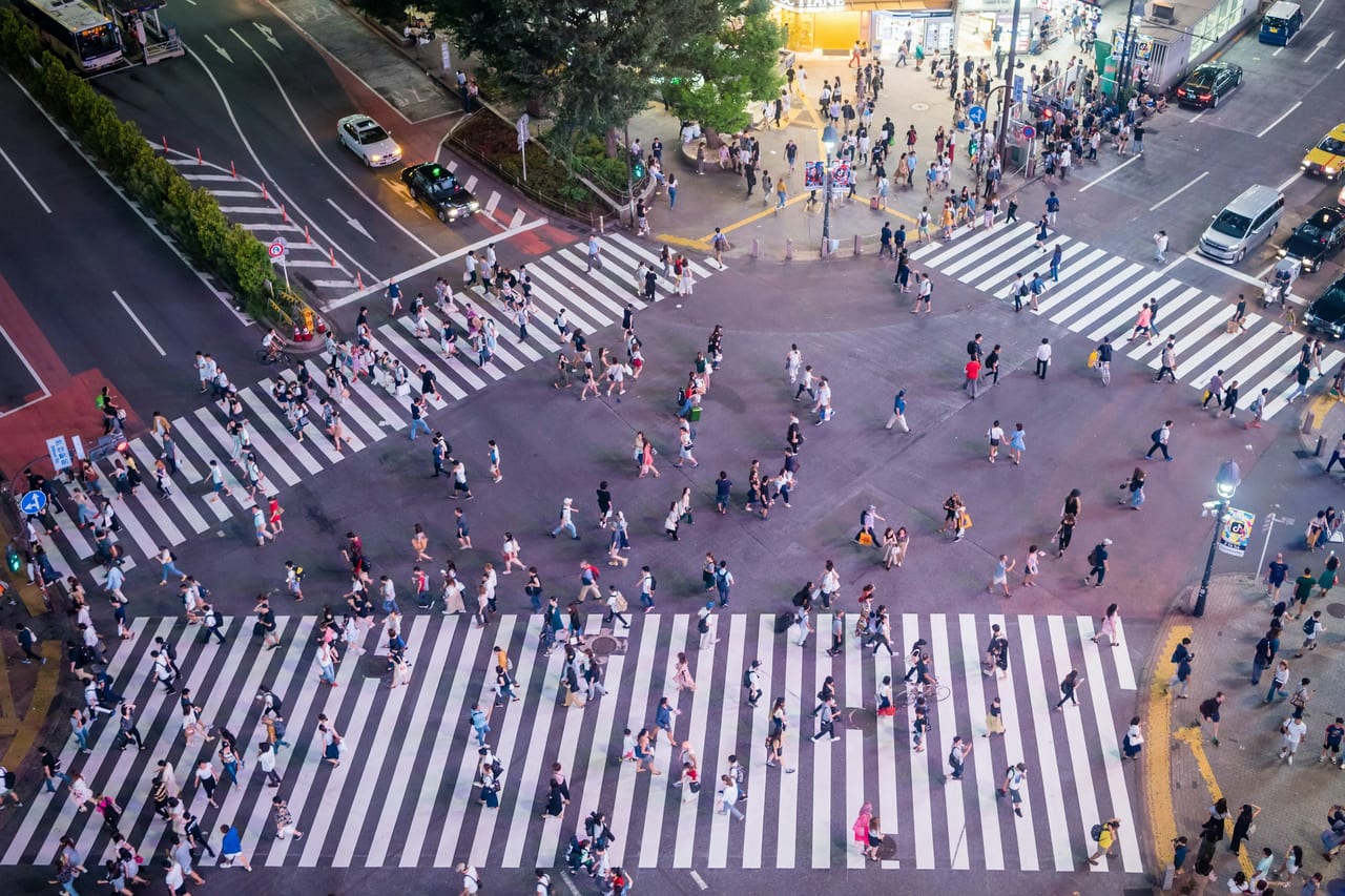 Crowds stream across the painted lanes of the Shibuya scramble crossing, seen from above in daylight.