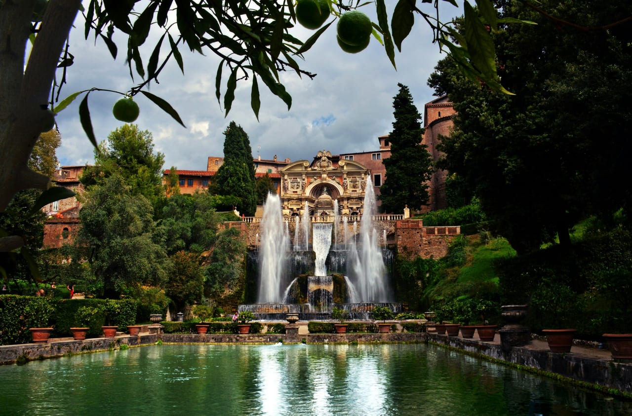 The Neptune and Organ fountains of Villa d'Este spill toward a still fish pond, framed by an orange-tree branch.