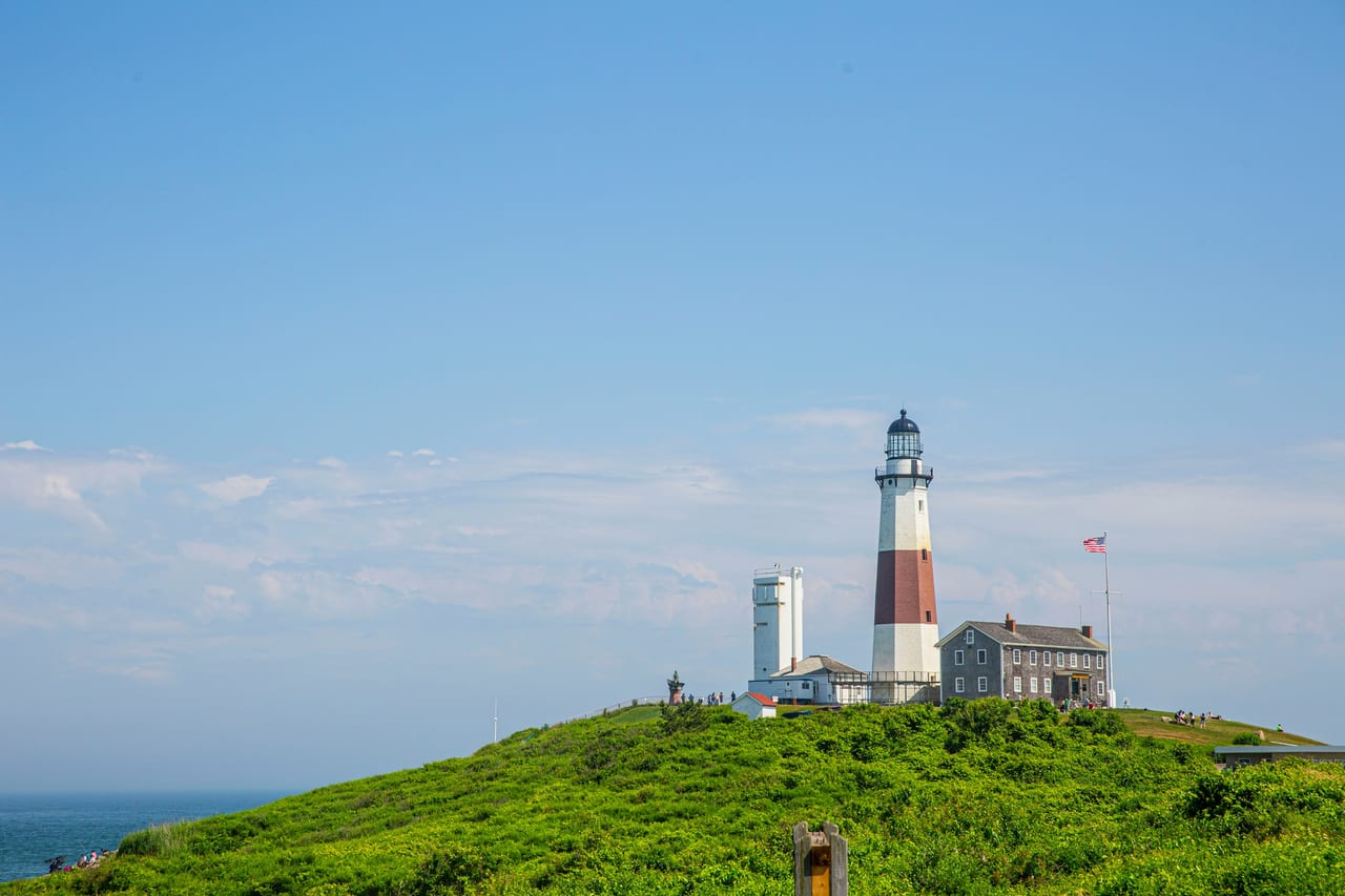 Montauk Point Lighthouse stands on a green hill beside the sea under a blue sky.