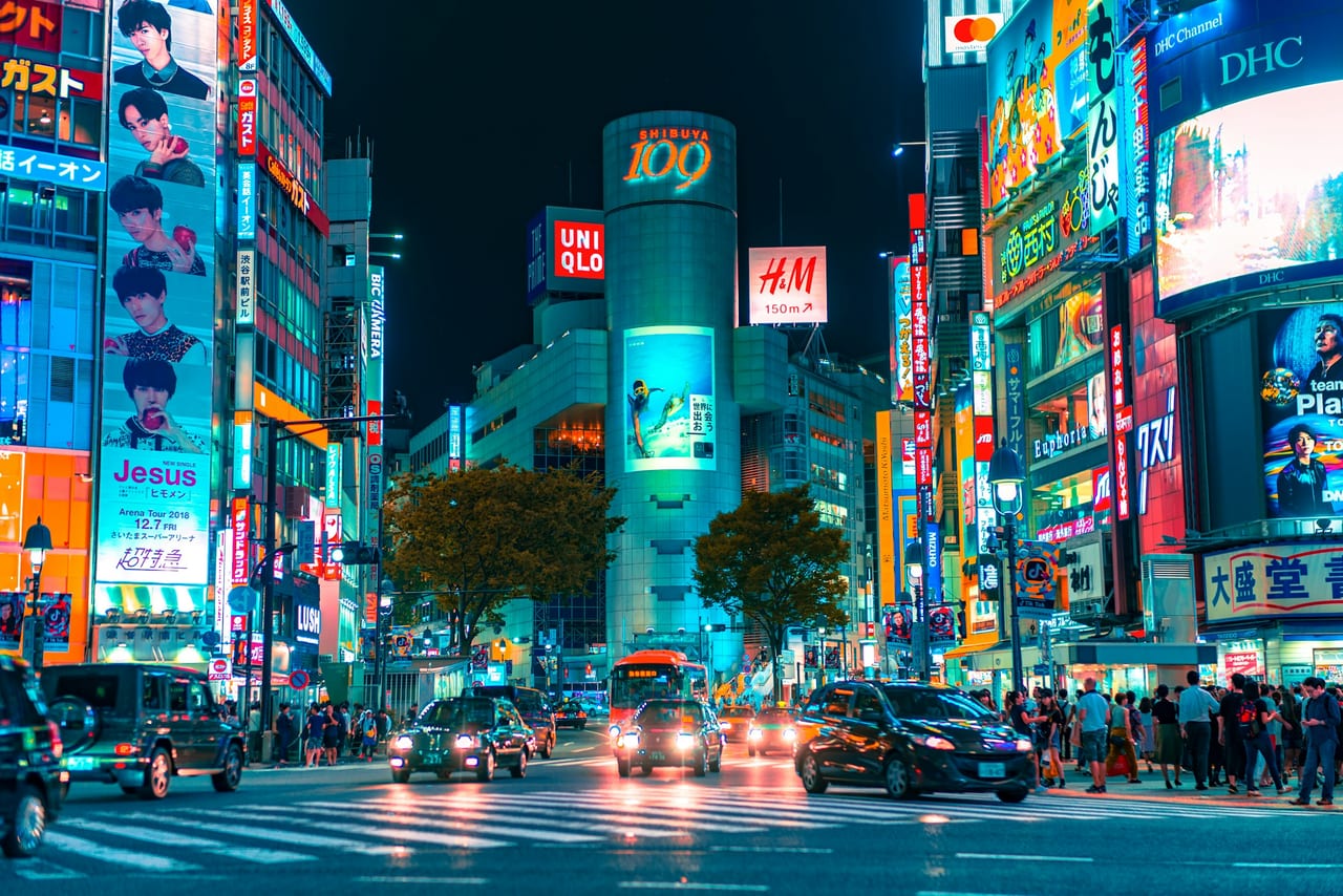 Neon billboards blaze over the Shibuya scramble crossing as traffic streaks past on a wet night.