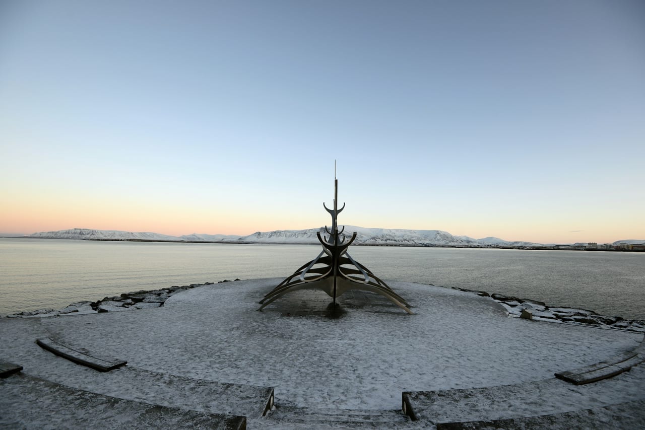 The Sun Voyager sculpture stands on Reykjavik's waterfront with snow-covered ground, calm water, and mountains in the distance.