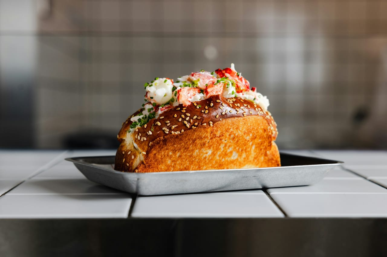 A toasted split-top brioche roll overflowing with chunks of lobster meat, chives, and sesame on a metal tray.