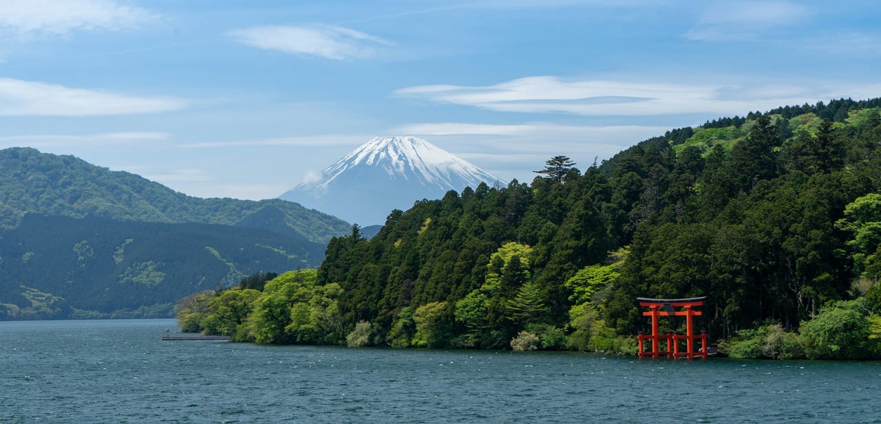 Snow-capped Mt. Fuji rises beyond Lake Ashi, with the red Hakone Shrine torii standing at the wooded shore.