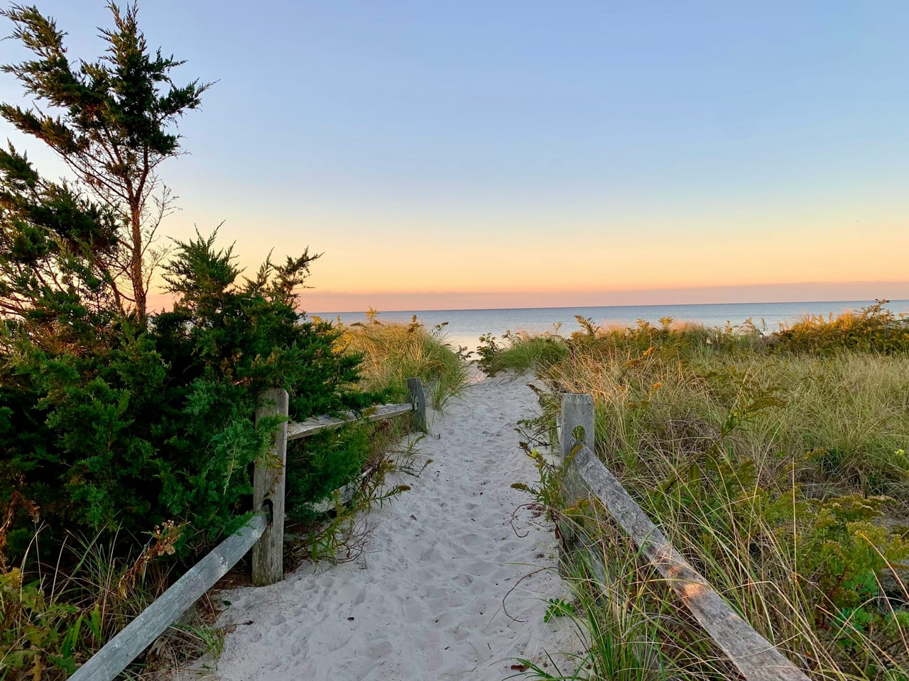 A sandy path flanked by beach grass and a wooden fence leads toward the ocean at dusk.