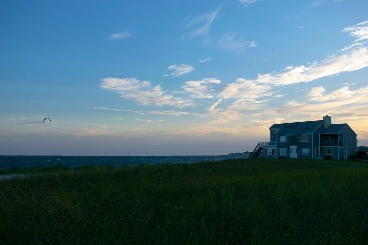 A shingled beach house sits on a grassy dune overlooking the ocean at dusk with a kite flying in the distance.