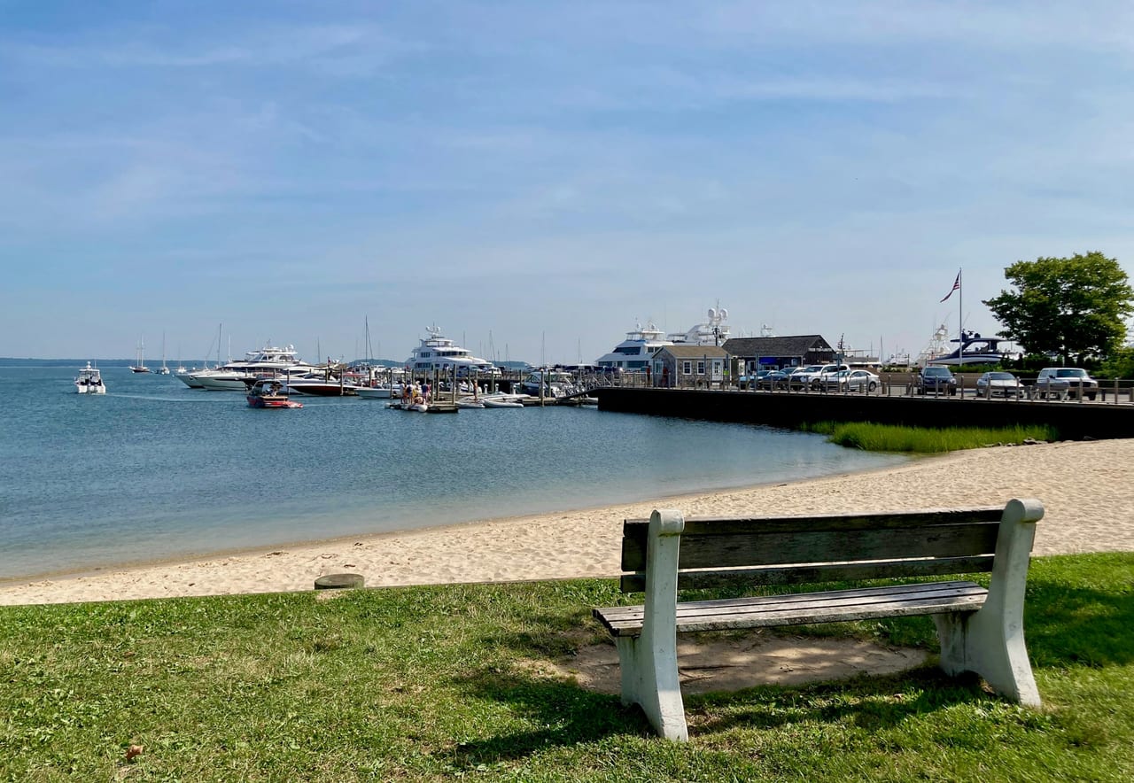 A wooden bench overlooks a sandy beach and harbor filled with docked boats under a clear daytime sky.