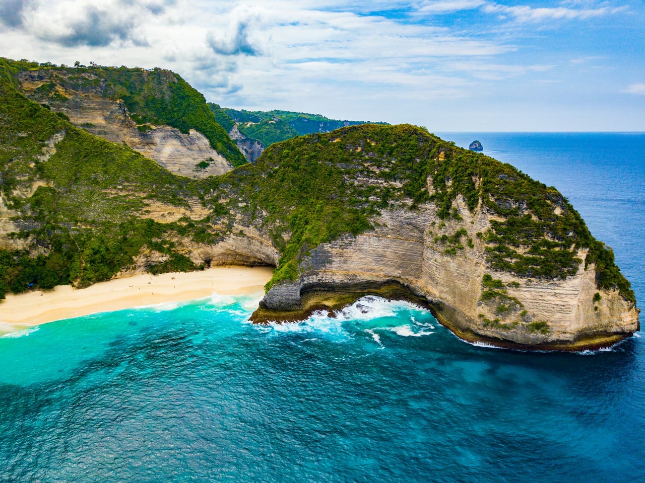 Aerial view of a steep green coastal cliff, turquoise water, and a sandy beach in Bali.