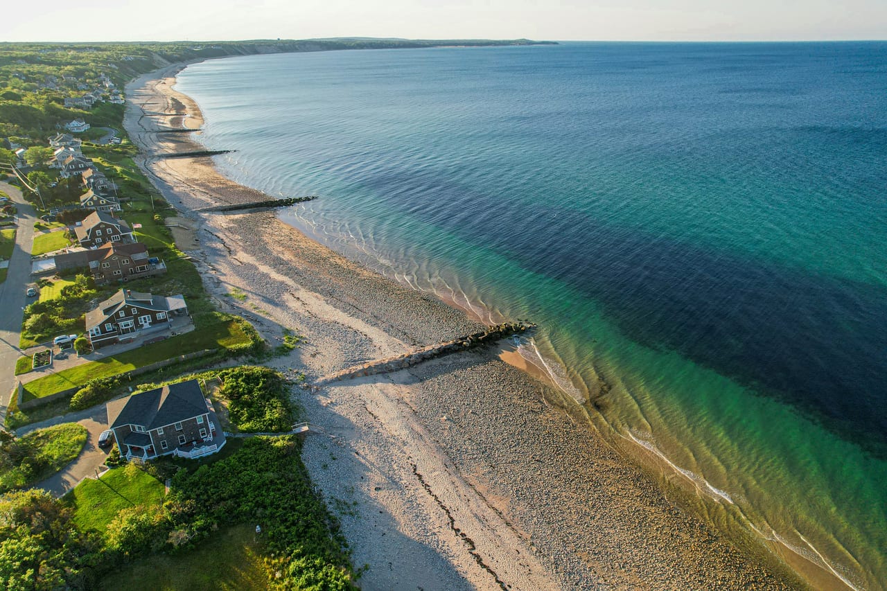 Aerial view of a Cape Cod shoreline with turquoise water, a stone jetty, and homes lining a sandy beach under bright sun.