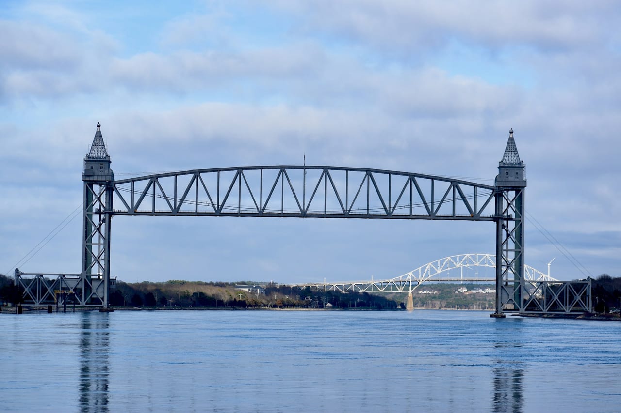 The Cape Cod Canal railroad lift bridge spans calm water with the Sagamore Bridge visible in the background under a pale sky.