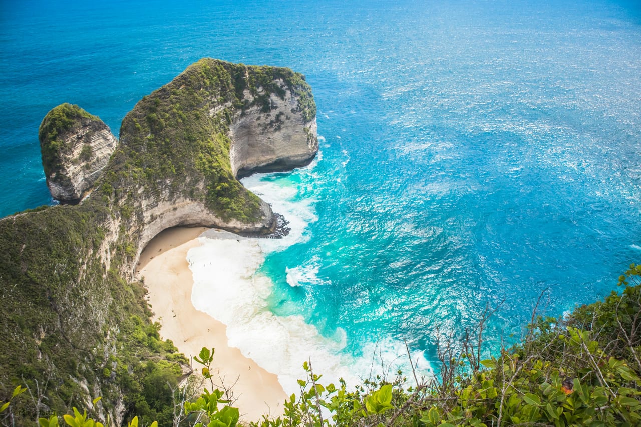 A white-sand beach curves beneath a steep green limestone headland surrounded by bright blue water.
